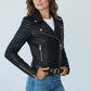 Scarlett Stitch black faux leather biker jacket with gold hardware, belted waist, and ribbed shoulders on a model.
