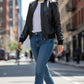 Scarlett Stitch women's black faux leather bomber jacket with ribbed collar, cuffs, and hem, worn by a model walking on a city street.