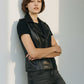 Scarlett Stitch black faux leather moto vest with lapel collar, zip accents, and buckle tabs. Model wears vest over black t-shirt and grey jeans.