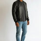 Scarlett Stitch black leather bomber jacket on a man wearing a grey t-shirt and blue jeans. Features ribbed collar, cuffs, and hem.