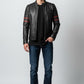 A man wearing a Scarlett Stitch black leather motorcycle jacket with red sleeve stripes, dark jeans, and white sneakers.