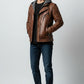 Scarlett Stitch men's brown distressed leather motorcycle jacket with a grey knit hood, worn with jeans and sneakers.