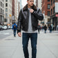 Scarlett Stitch Men's black faux leather bomber jacket with dark brown shearling collar, zip front, and flap pockets.