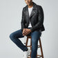 Man wearing a black Scarlett Stitch leather biker jacket with silver hardware, white t-shirt, and blue jeans on a stool