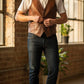Scarlett Stitch Men's distressed brown leather vest with V-neck, button front, and flap pockets, worn over a white t-shirt.