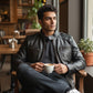 Scarlett Stitch black faux leather bomber jacket with stand collar and zippered pockets, worn by a man sitting in a cafe.