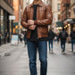 Scarlett Stitch Men's vintage distressed brown genuine leather bomber jacket with ribbed collar, cuffs, and hem. Model wears it with a black t-shirt and blue jeans.
